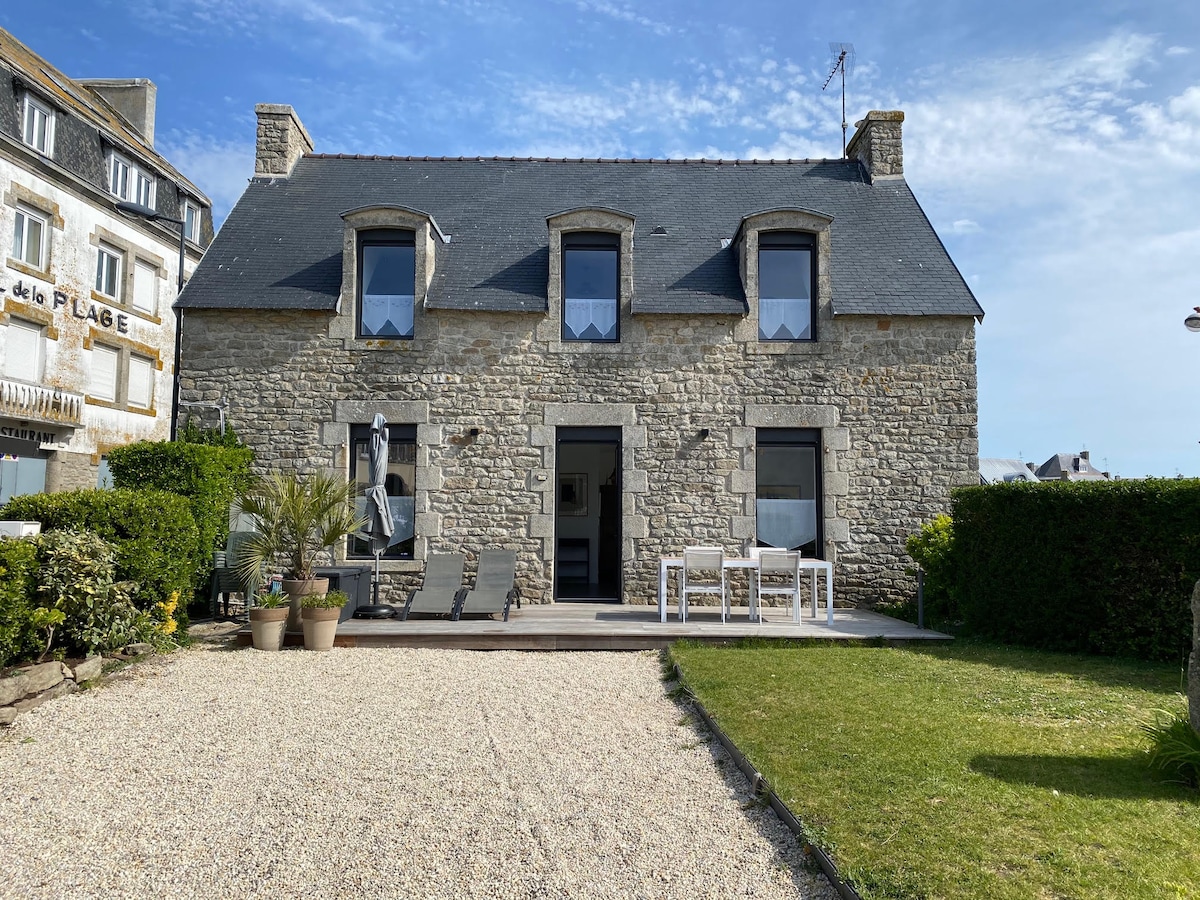 A traditional stone house with a slate roof is set in a landscaped garden, featuring a gravel pathway and green lawn. The front porch includes a table and chairs, inviting outdoor gatherings. Large windows allow natural light to enter, enhancing the home's charm.