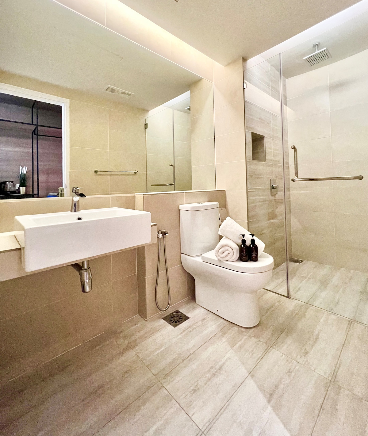 The bathroom features a glass shower enclosure and a modern toilet. A wall-mounted sink with a sleek design is located beside it. Toiletries are arranged neatly on the countertop, while light-colored tiles enhance the spacious and bright atmosphere.