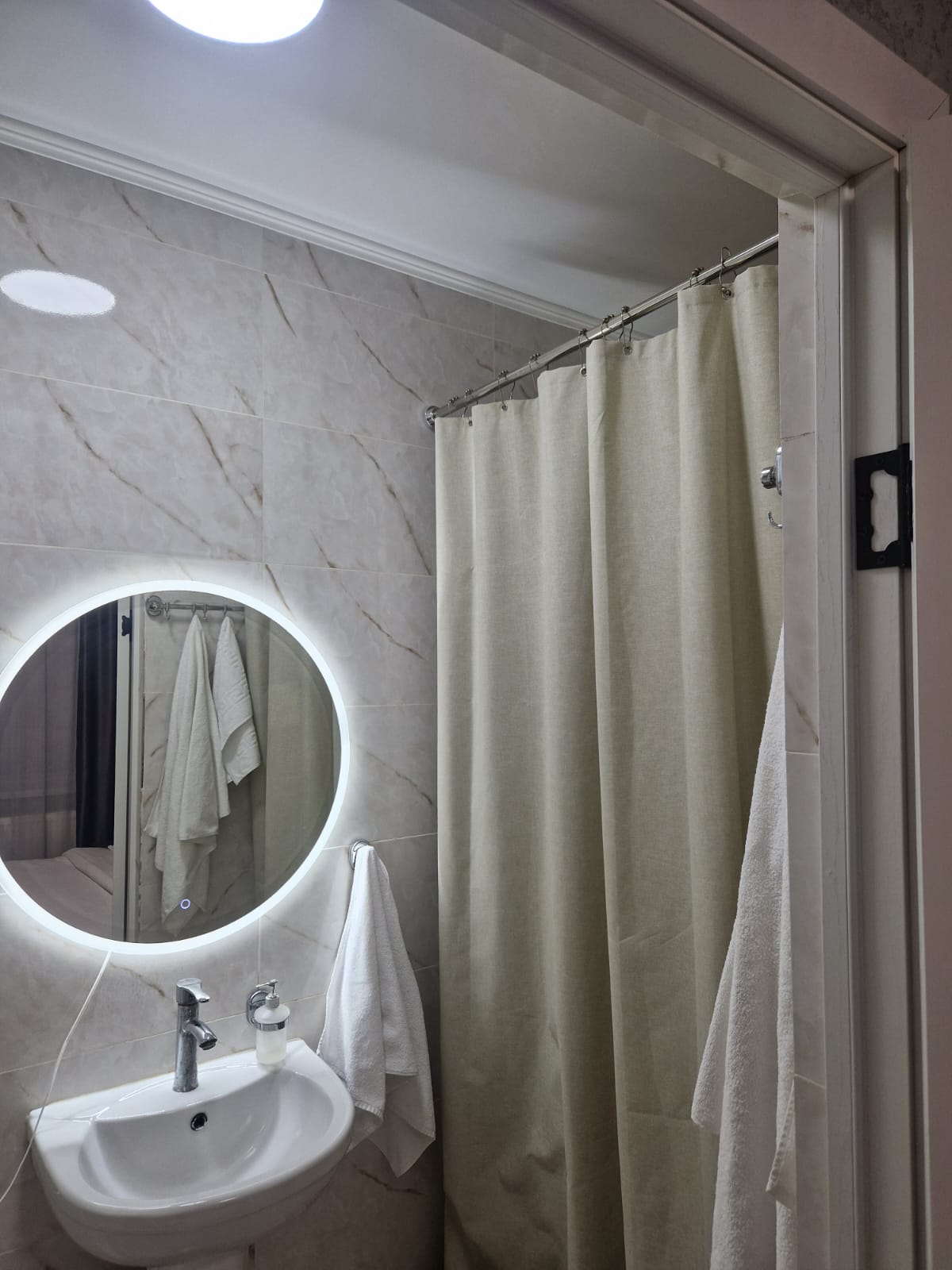 A modern bathroom features light-colored marble walls and a circular mirror with built-in lighting. A compact sink is positioned beneath the mirror, while a shower area is partially enclosed by a neutral-toned curtain. A set of white towels is neatly hung nearby.