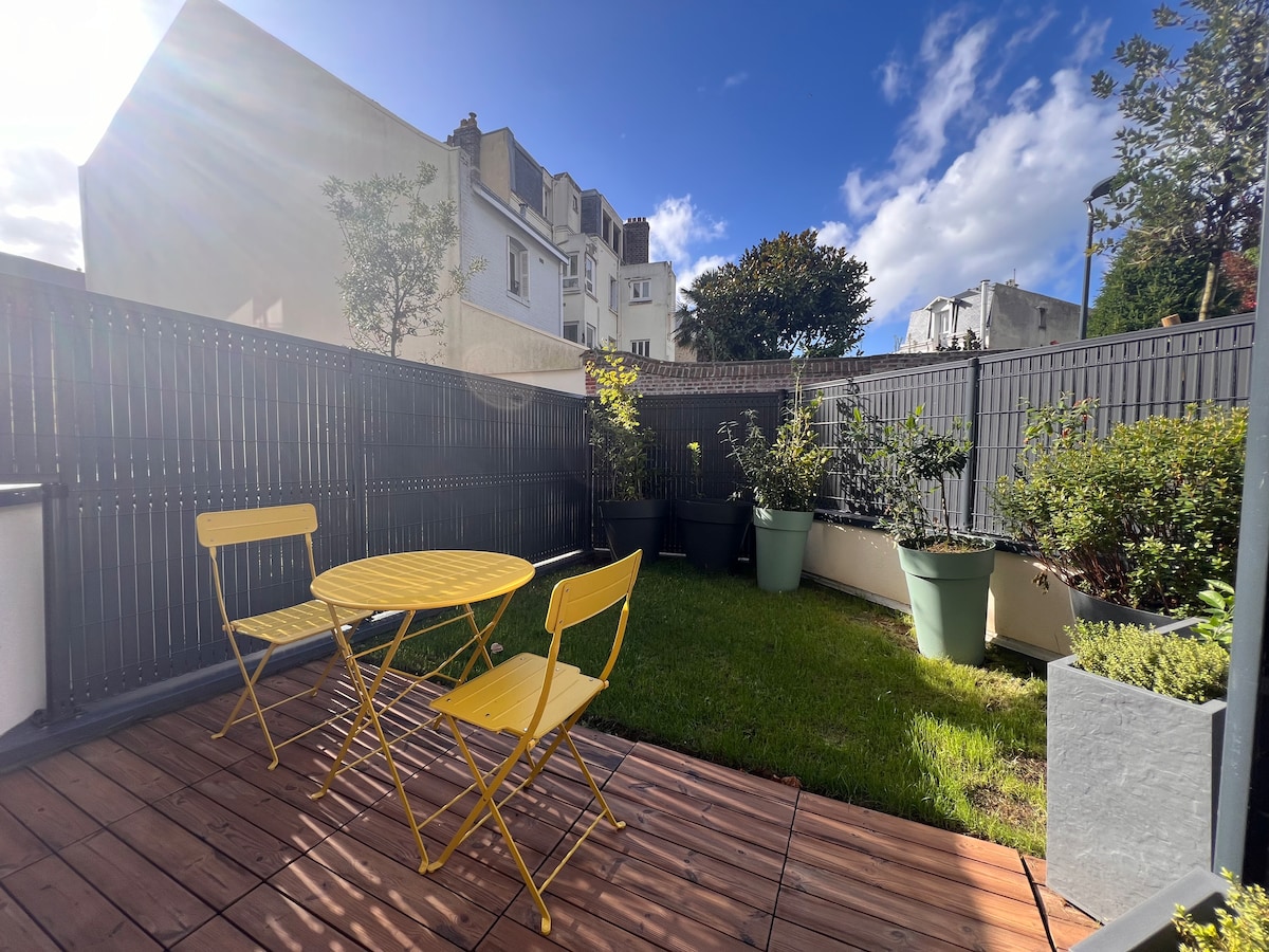 A private terrace is presented, featuring a wooden deck and lush grass. Two yellow metal chairs and a round table are set against a backdrop of planters filled with greenery. The sky is partly cloudy, allowing natural light to illuminate the space.