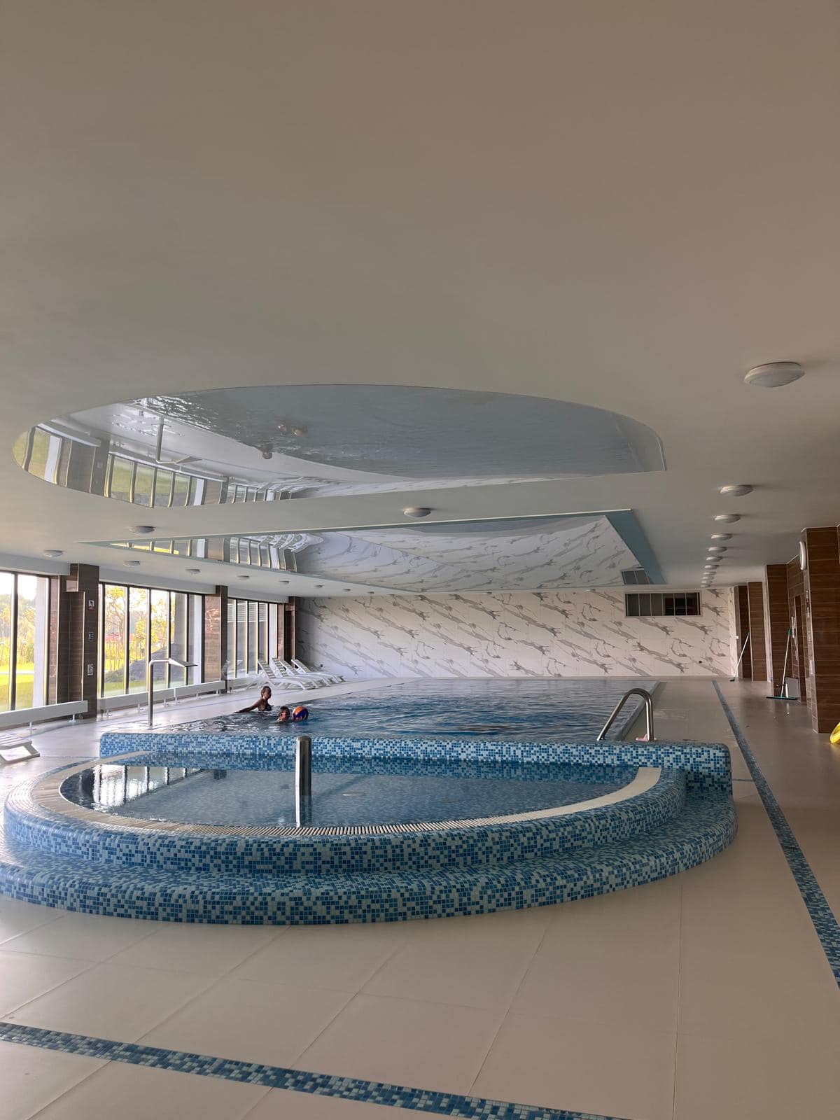 A clean and modern wellness area features a jacuzzi surrounded by decorative blue tiles. Large windows allow natural light to fill the space, with views of the outdoors visible through the glass. Sleek lounge chairs are positioned nearby for relaxation.