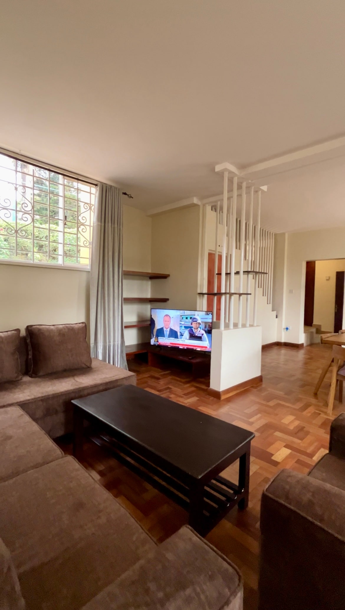 A living area is featured with a dark wooden coffee table positioned in front of a brown sectional sofa. A television is placed on the floor, displaying a program. Large windows allow natural light to fill the room, complemented by neutral-toned curtains.