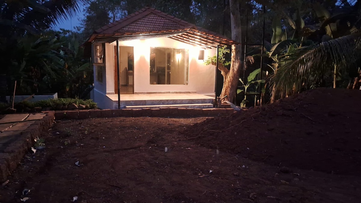 A tranquil cottage is illuminated softly at dusk, showcasing large glass doors that provide an inviting entrance. Surrounding greenery enhances the natural setting, while a spacious area in front offers potential for outdoor activities. The rustic roof adds charm to the serene landscape.