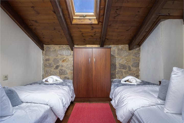 ห้องนอนใต้หลังคาสว่างและอบอุ่นมีเตียงเดี่ยวสองเตียงใต้เพดานคานไม้ที่มีช่องแสง เสน่ห์แบบชนบทและแสงธรรมชาติทำให้เป็นที่พักที่น่าผ่อนคลาย