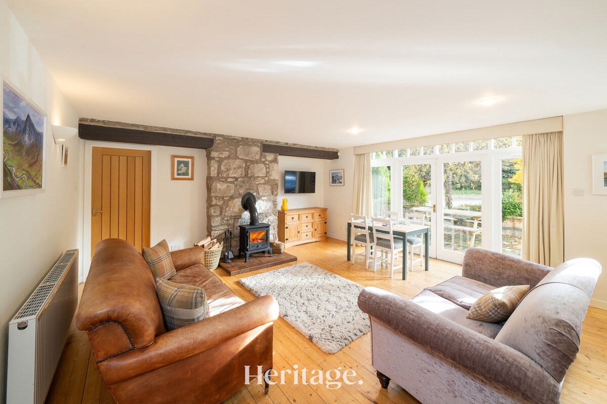 An open-plan living area features a cozy seating arrangement with a brown leather sofa and a light-colored armchair. A log-burning stove is positioned against a stone wall, and large glass doors provide access to an outdoor dining area. A wooden dining table is visible in the background.