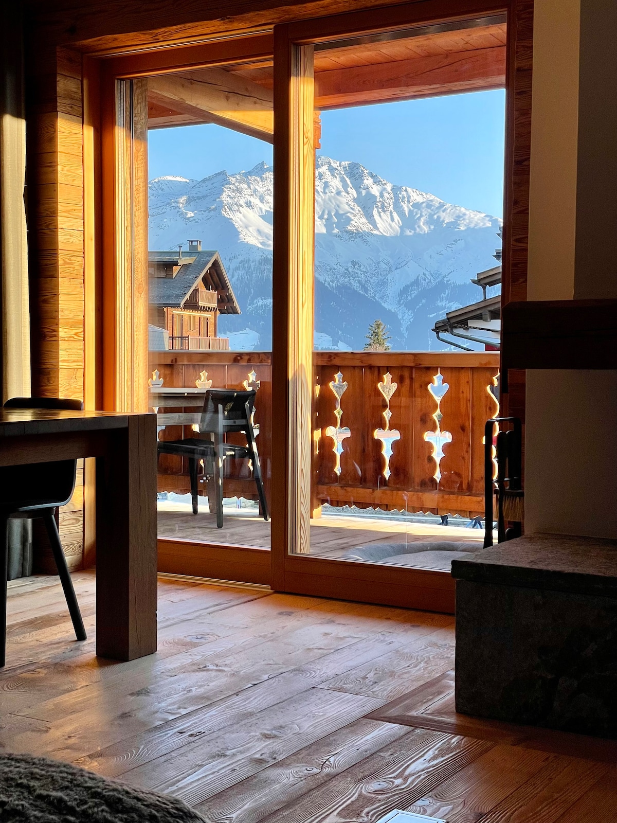 A glass door opens to a balcony, revealing a stunning view of snow-capped mountains. Natural wood framing complements the warm tones of the interior, with a rustic wooden floor and furniture partially visible in the foreground.