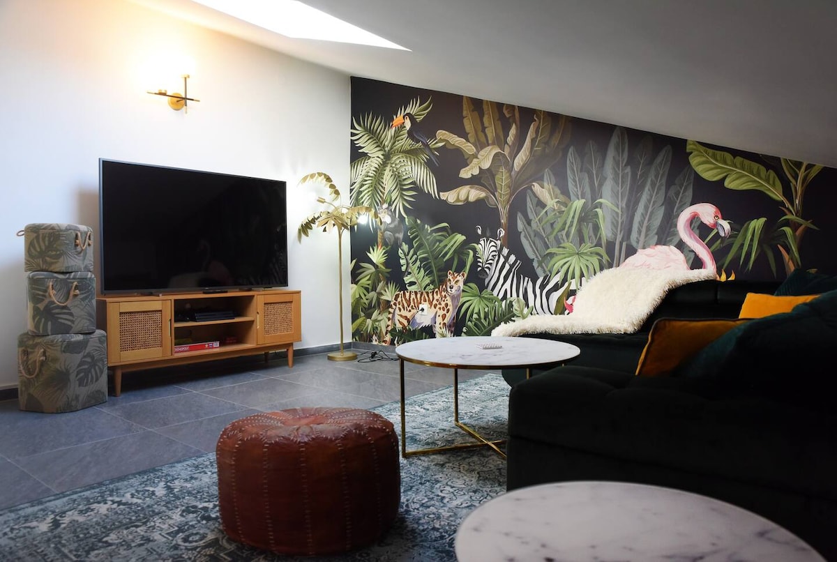 A stylish living area features a large flat-screen television positioned on a mid-century modern cabinet. A tropical mural with palm leaves and animals, including a flamingo, serves as a vibrant backdrop. Two circular coffee tables and a plush ottoman are present, enhancing the cozy atmosphere.