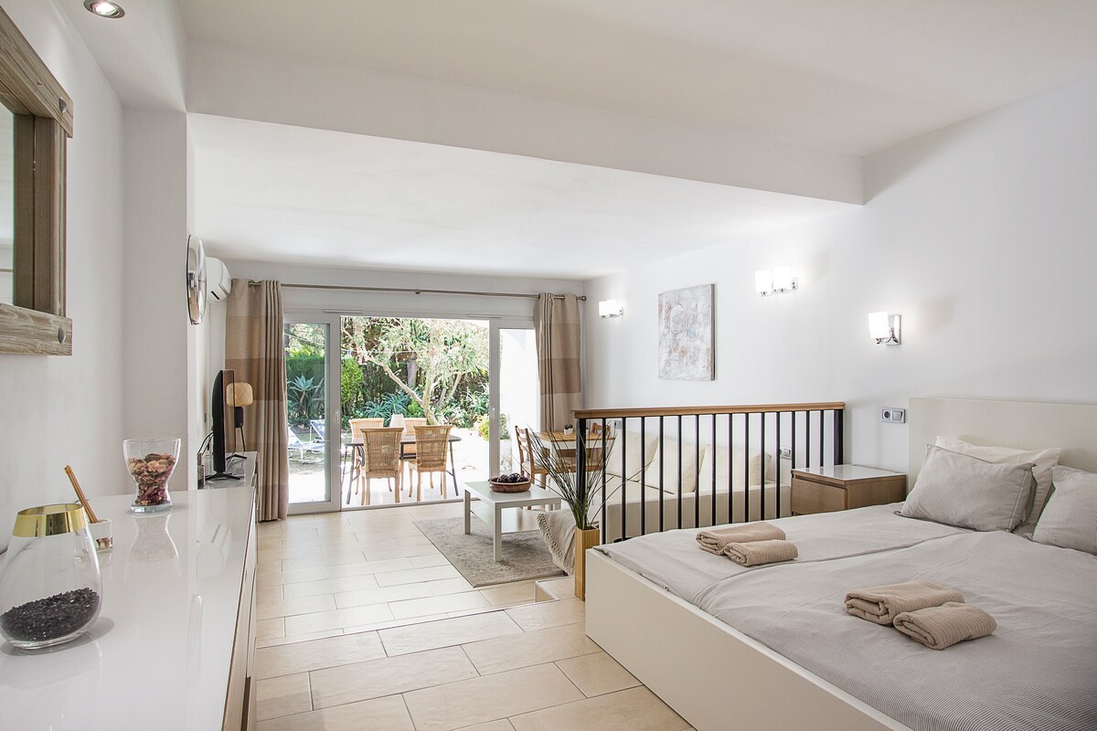 A spacious studio layout is presented, featuring a comfortable double bed with light linens, positioned adjacent to a seating area. Large glass doors open to a terrace with outdoor dining furniture, providing views of the surrounding greenery. Soft lighting enhances the inviting atmosphere.