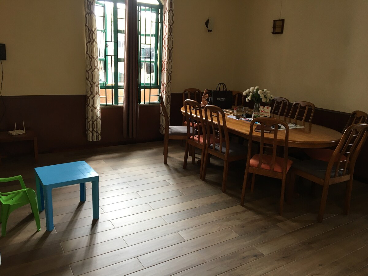 A spacious dining area features a large wooden table surrounded by multiple chairs, suitable for gatherings. Two windows allow natural light, adorned with curtains. A small blue table and a green chair are positioned on the floor, adding a playful touch to the space.