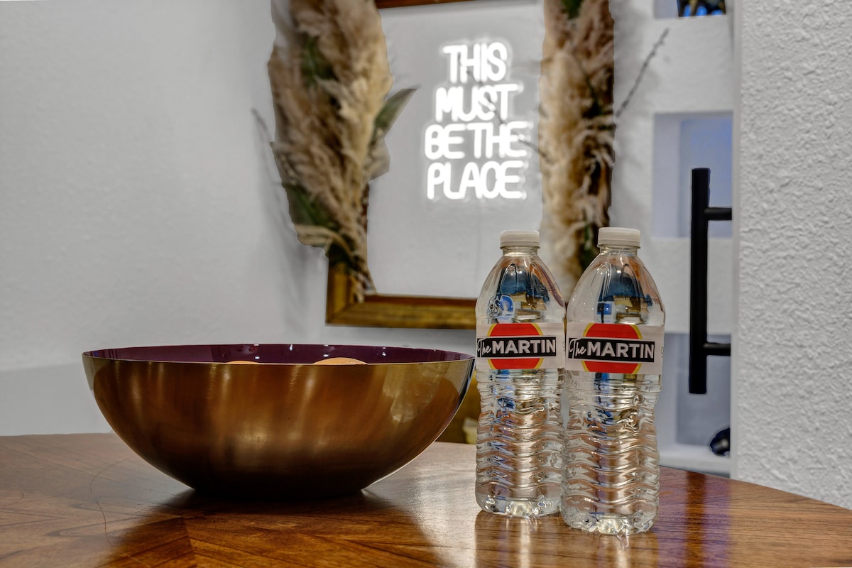 A wooden table is set with a metallic bowl and two labeled water bottles beside it. A lighted sign in the background reads 'THIS MUST BE THE PLACE,' framed by decorative elements that add a touch of warmth to the space.