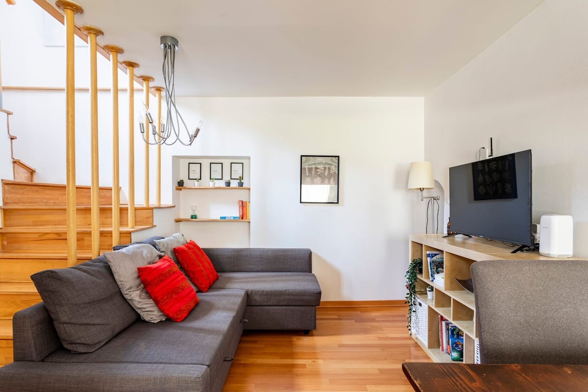 A spacious living area is present, featuring a gray sectional sofa adorned with red cushions. A flat-screen TV is positioned on a wooden shelf, while a dining table and chairs are visible nearby. Natural light fills the room, complemented by a modern light fixture and wooden stairs leading to the upper level.