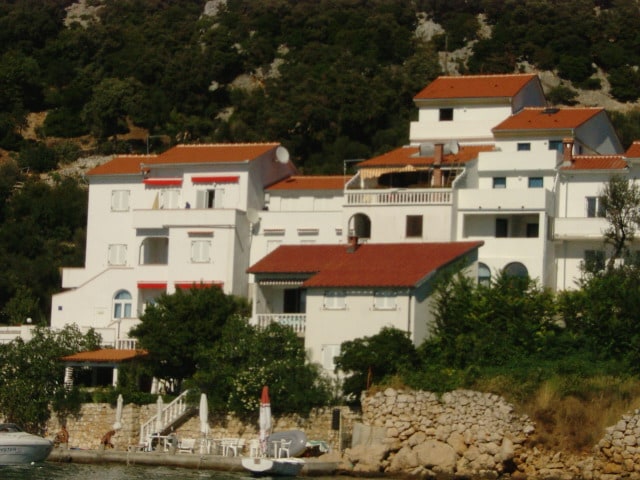 A multi-story building with a combination of white and red accents overlooks the water. The structure features balconies and terraced areas, surrounded by lush greenery and stone paths leading to the shoreline. A small boat is visible near the dock area.