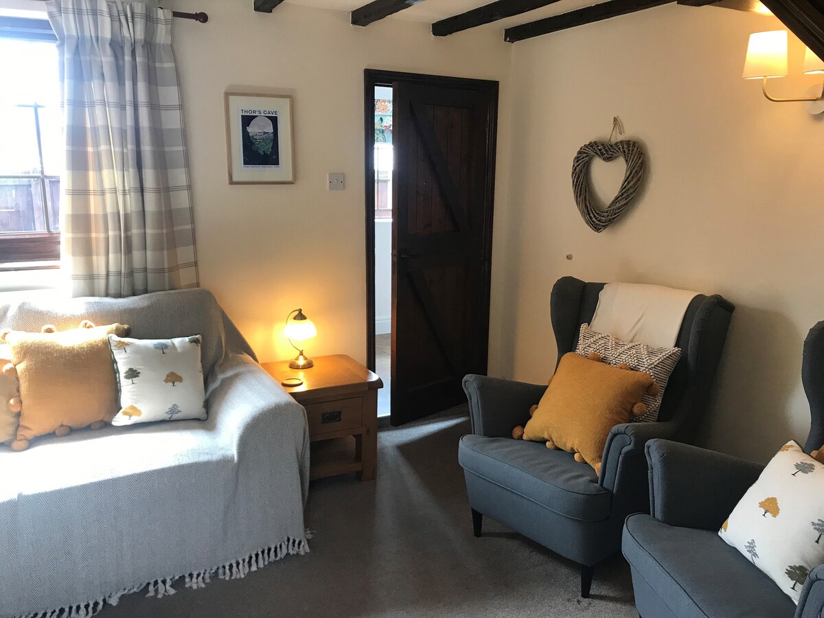 A cozy living room features soft furnishings in muted tones. A light gray sofa is placed beside a wooden side table with a warm lamp. Two comfortable armchairs with decorative cushions provide additional seating. Exposed wooden beams complement the rustic charm of the space.