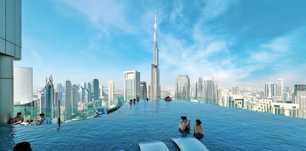 A rooftop infinity pool is visible, reflecting the skyline of Downtown Dubai against a clear blue sky. Guests can be seen enjoying the water, while the Burj Khalifa towers in the background, surrounded by modern skyscrapers.
