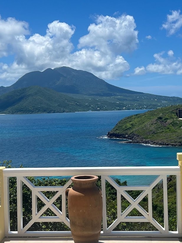 A scenic view is captured from a terrace, showcasing a sweeping vista of the ocean and lush mountains in the distance. A clay pot is positioned prominently in the foreground, adding a touch of character to the serene landscape.