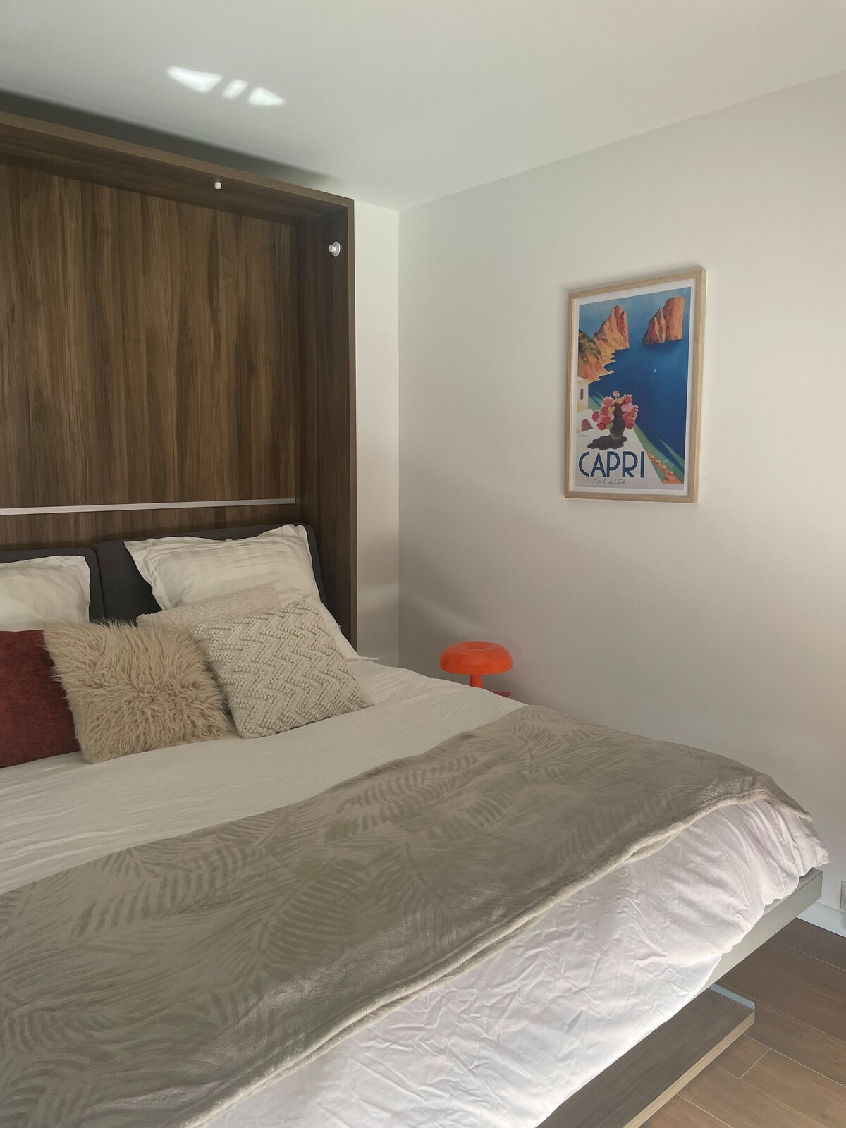 A cozy sleeping area is featured, showcasing a spacious bed with decorative pillows and a soft blanket. A wooden wall unit offers a modern touch, while a vibrant artwork depicting 'Capri' adds a pop of color. Natural light highlights the warm tones of the room.