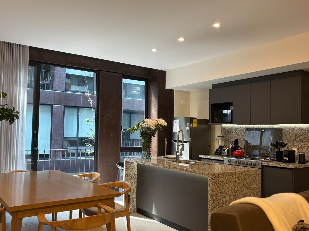 A modern kitchen is complemented by a spacious dining area, featuring a wooden table and several light-colored chairs. A large window brightens the space, offering views of greenery. The kitchen showcases sleek countertops with various appliances, providing a functional and inviting atmosphere.