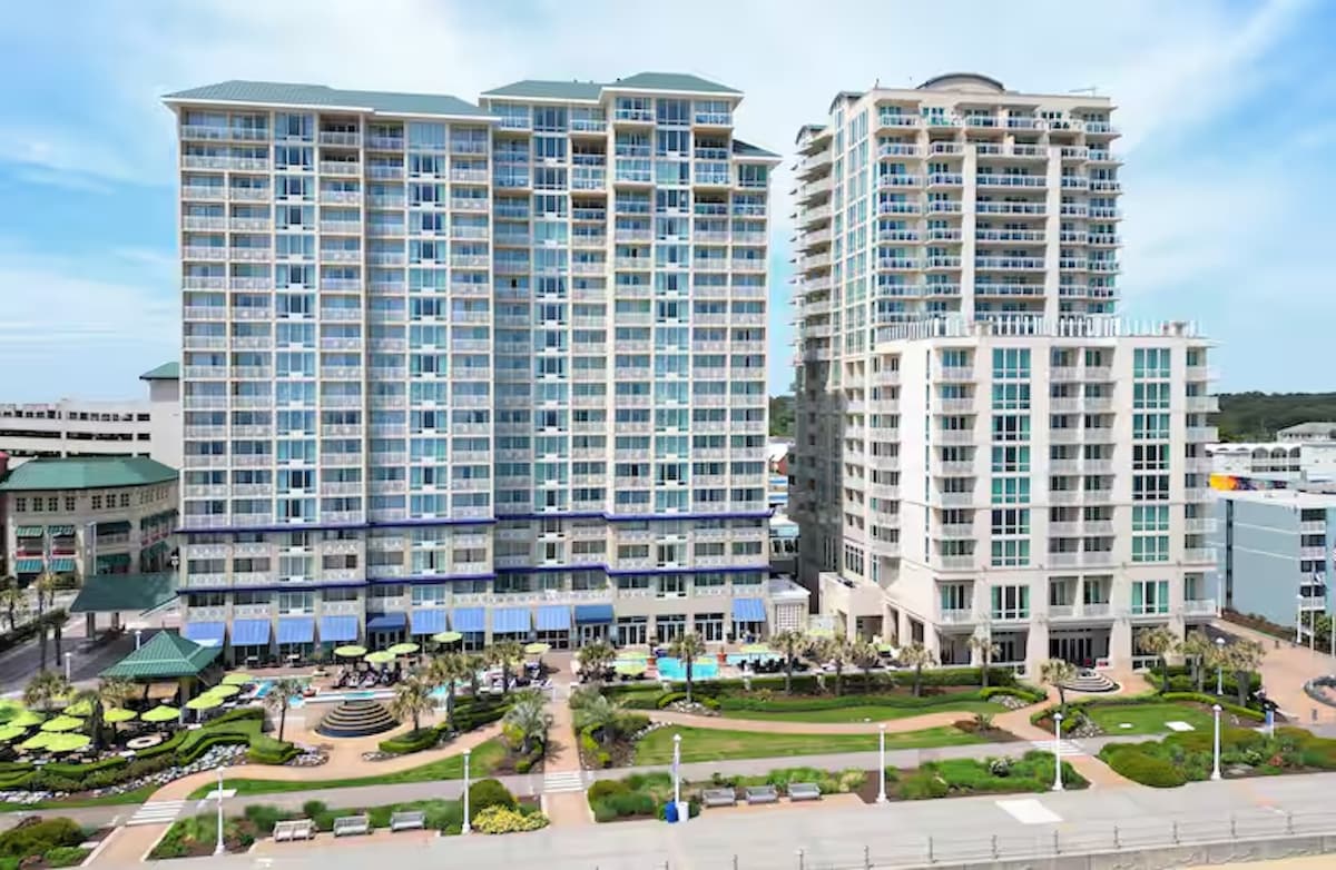 The Oceanaire Resort is showcased from an aerial perspective, highlighting its modern architecture. Expansive glass balconies offer ocean views, while lush landscaping surrounds the building. A series of pools and seating areas can be seen below, emphasizing the resort's inviting outdoor spaces.