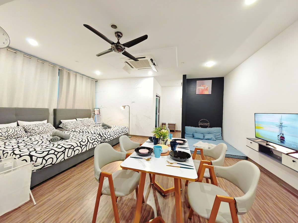 An open studio space features two queen-sized beds adorned with patterned bedding, complemented by a cozy sofa in the corner. A dining table with four chairs is set for meals, while a wall-mounted TV and decorative art enhance the contemporary design.
