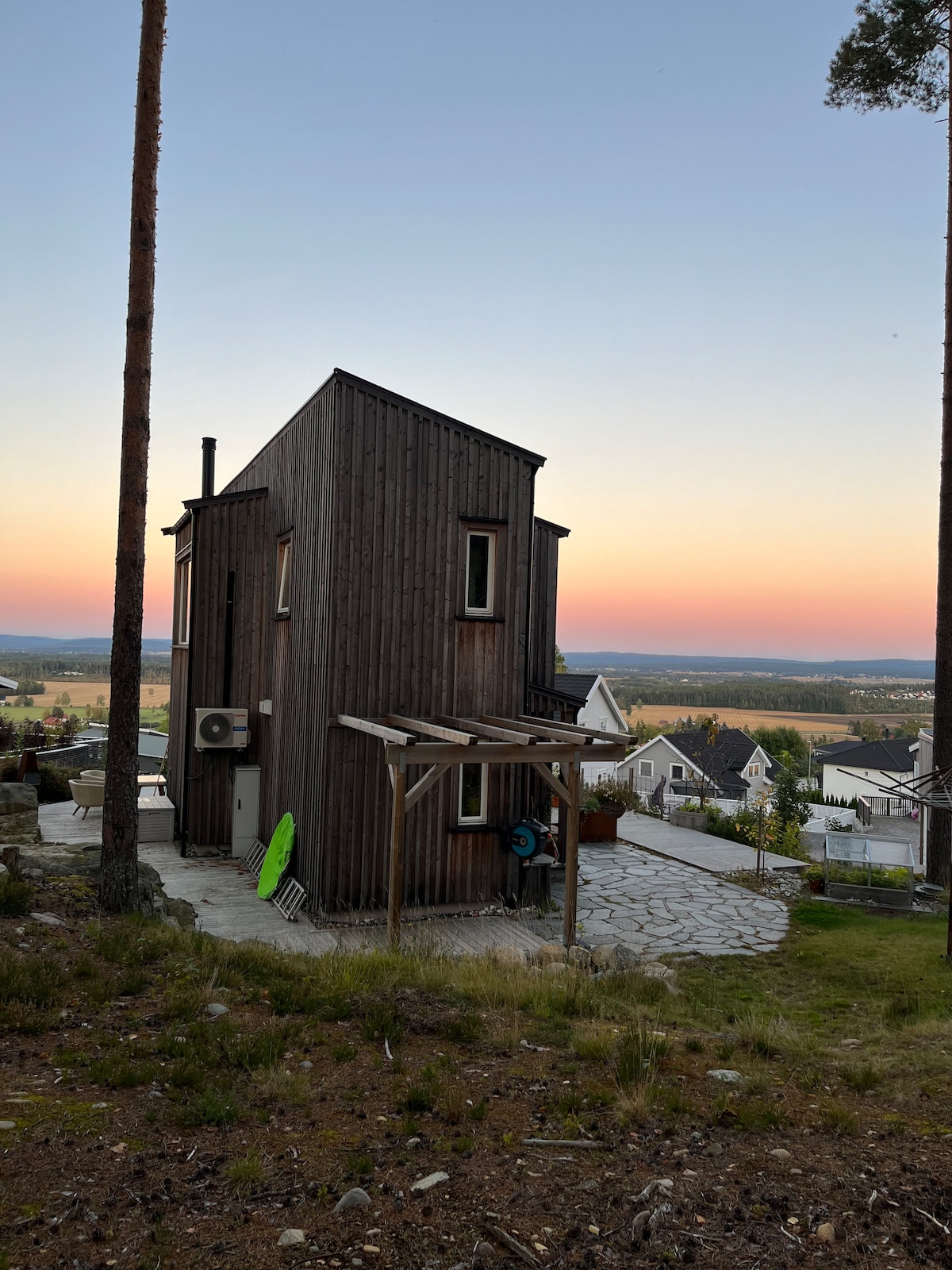 A modern wooden structure is positioned on a gentle slope, showcasing a unique slanted design. The exterior features a combination of dark wood and large windows. A stone patio area is visible in the foreground, with a surfboard leaning against the side of the building, and a colorful sunset in the background.