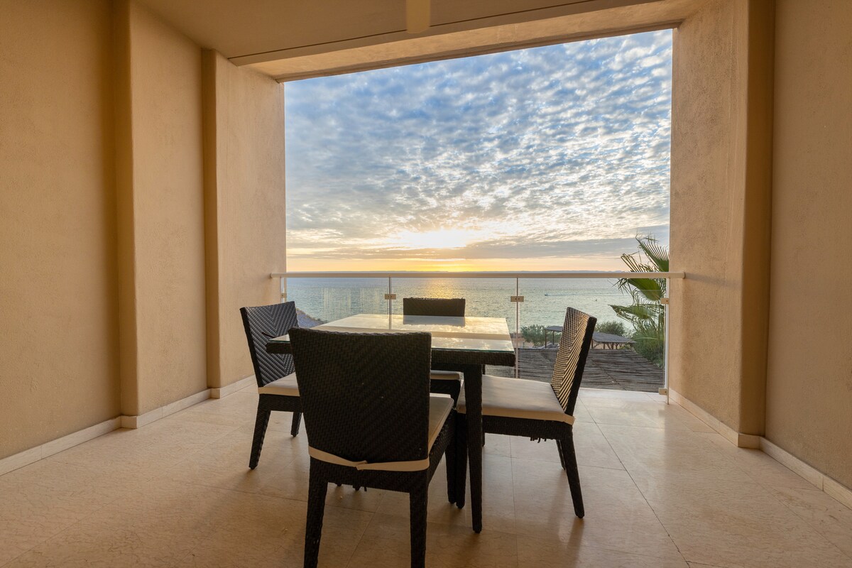 An indoor terrace area features a glass-top dining table surrounded by four black woven chairs. Large openings offer panoramic views of the ocean and sky, with soft clouds illuminated by the setting sun.
