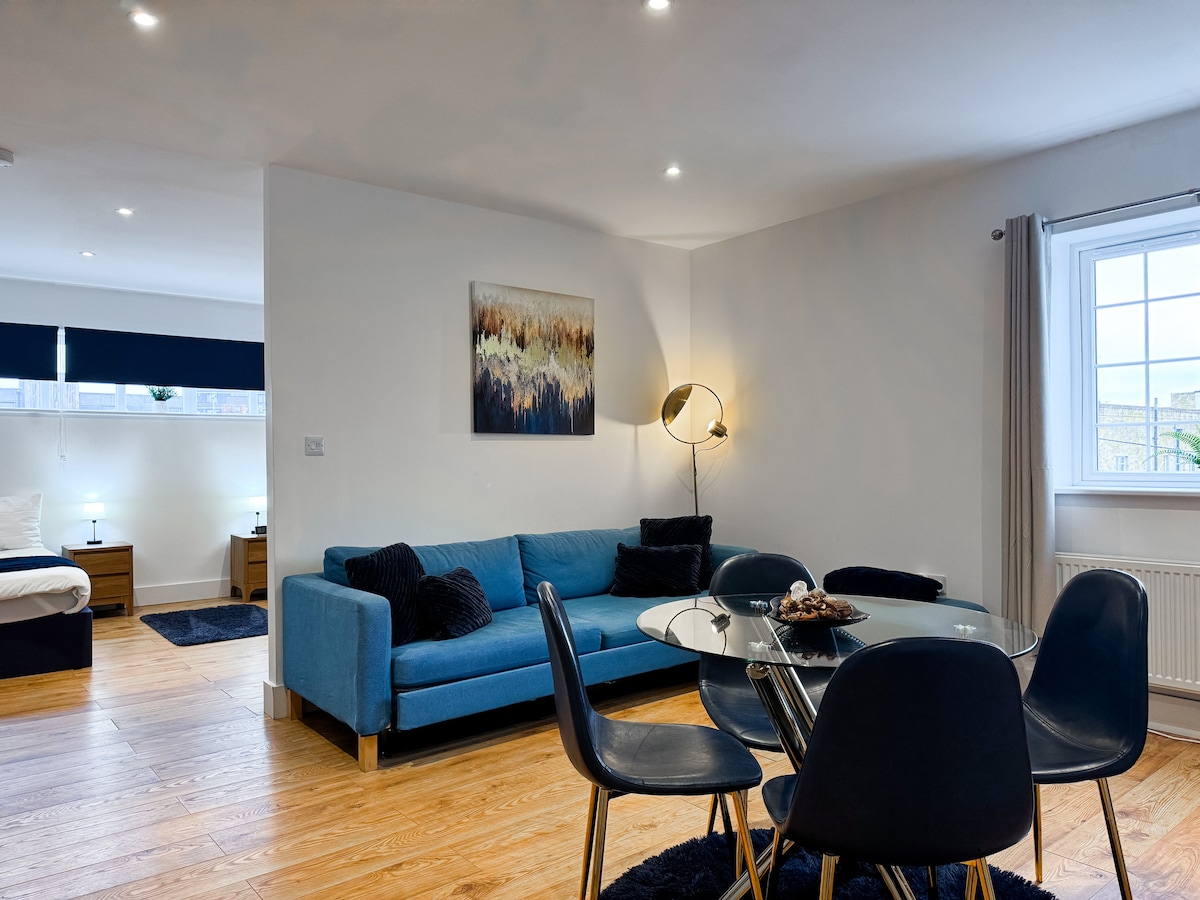 A stylish living area features a blue sofa paired with four black dining chairs around a glass table. A piece of abstract wall art adds visual interest. Natural light enters through a window, illuminating the warm wood flooring and creating a comfortable atmosphere.