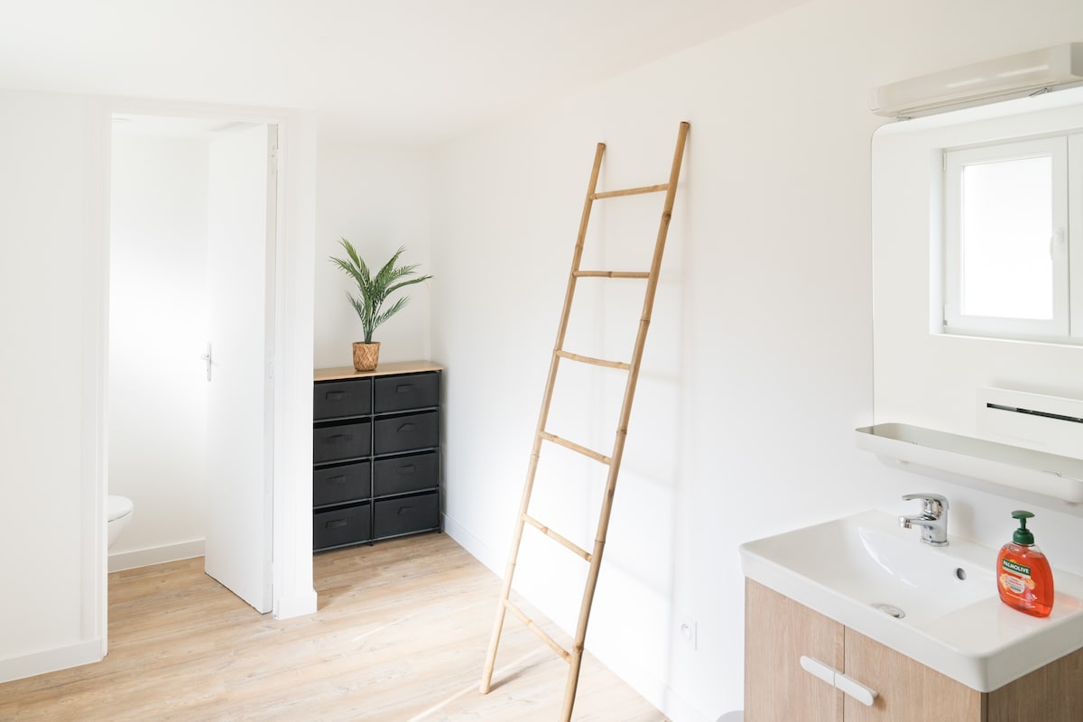 A bright and airy room features light wooden flooring and white walls. A decorative ladder leans against the wall, while a small potted plant adds a touch of greenery. A dark dresser is positioned adjacent to a window, which allows natural light to fill the space. A sink is also visible.