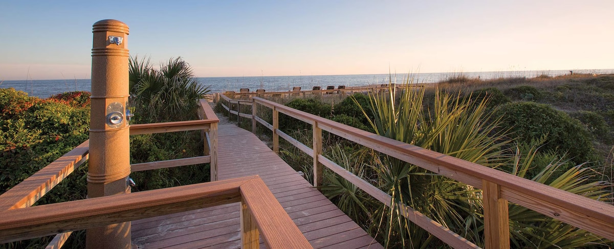 A wooden boardwalk leads to the beach, surrounded by lush greenery and palm plants. The pathway is illuminated by the soft light of a setting sun, offering a view of the ocean in the distance.