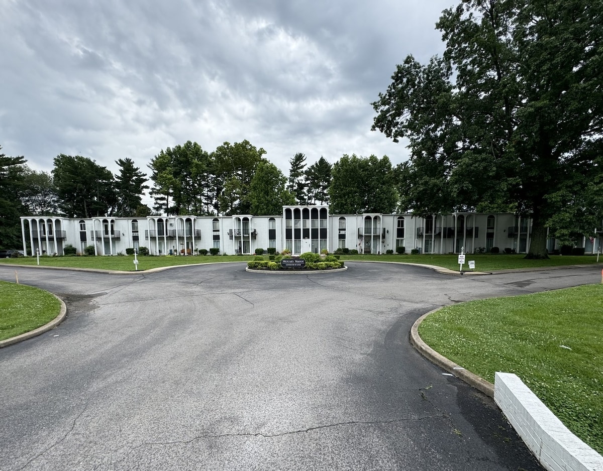 The entrance to a condo complex features a circular driveway surrounded by well-maintained green lawns and trees. The white façade of the building is accented by large, symmetrical arches that frame the entrance, creating an inviting and open appearance.