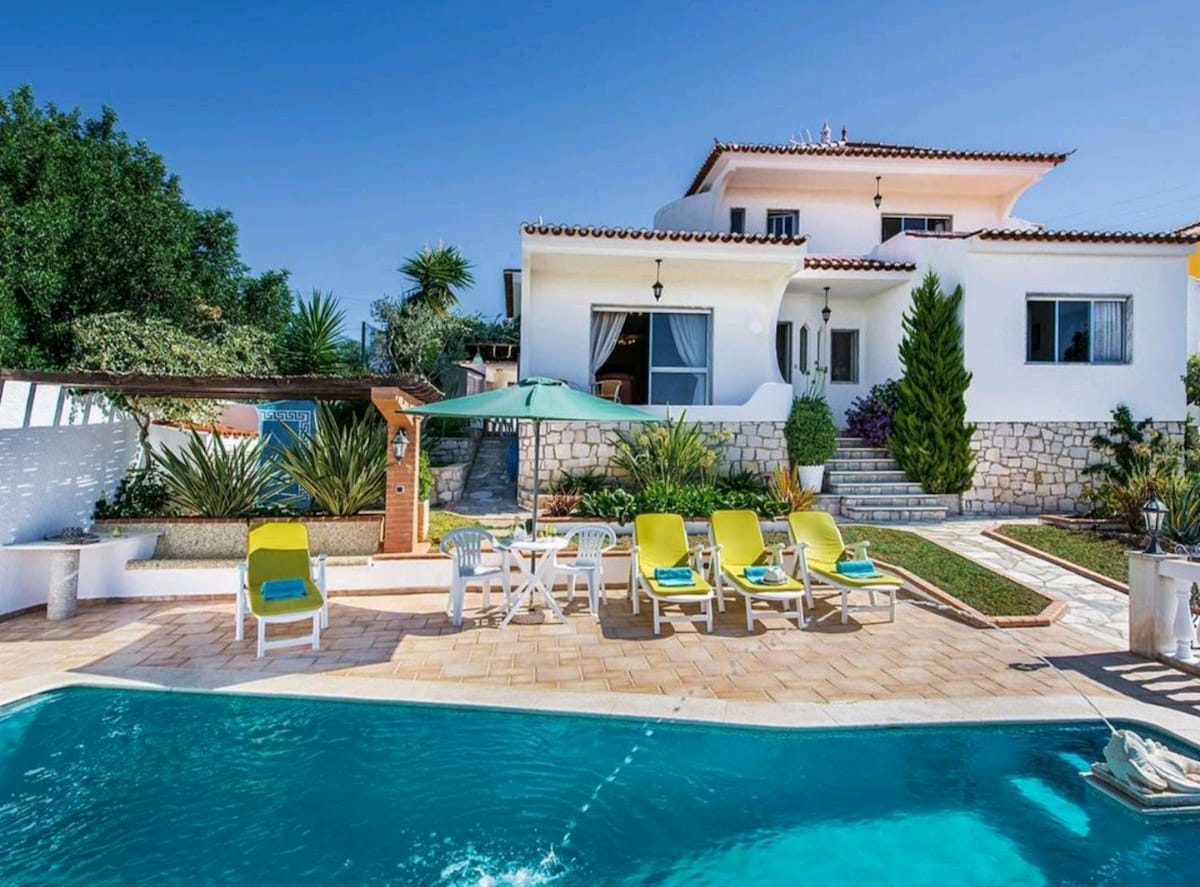 The exterior of the villa showcases a private pool surrounded by four bright yellow loungers and a shaded dining area. Lush greenery and decorative plants frame the property, while the two-story villa features large windows and a welcoming entrance leading to the garden.