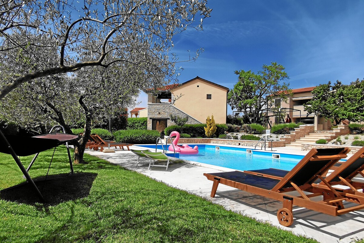A serene garden area features a large pool surrounded by lounge chairs. A pink flamingo float is visible in the water, complemented by lush greenery and trees. The setting includes a hammock nearby and the architecture of the surrounding houses can be seen in the background.