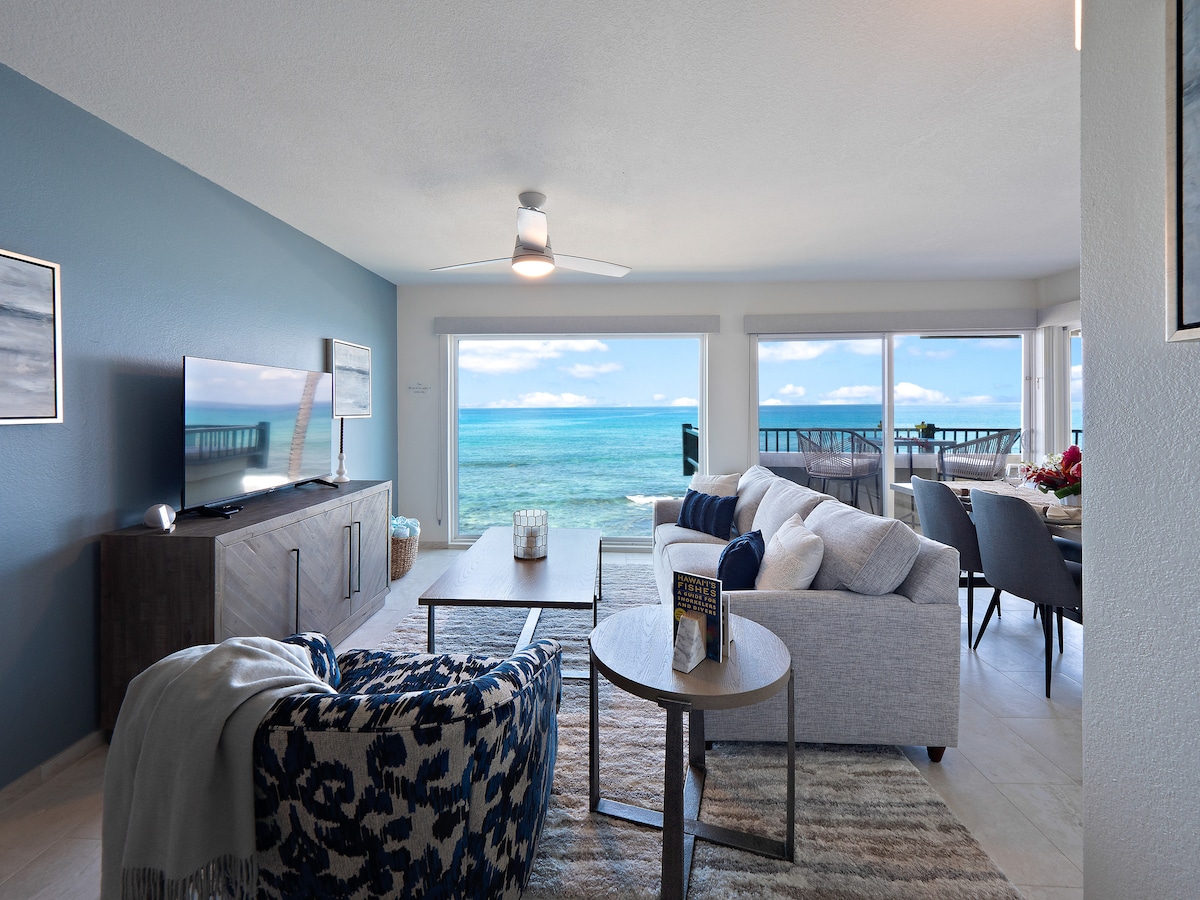 An open-concept living area is highlighted by large windows offering ocean views. A neutral sofa is complemented by patterned chairs and a coffee table. A television is positioned on a contemporary media console, enhancing the comfortable atmosphere of the space.