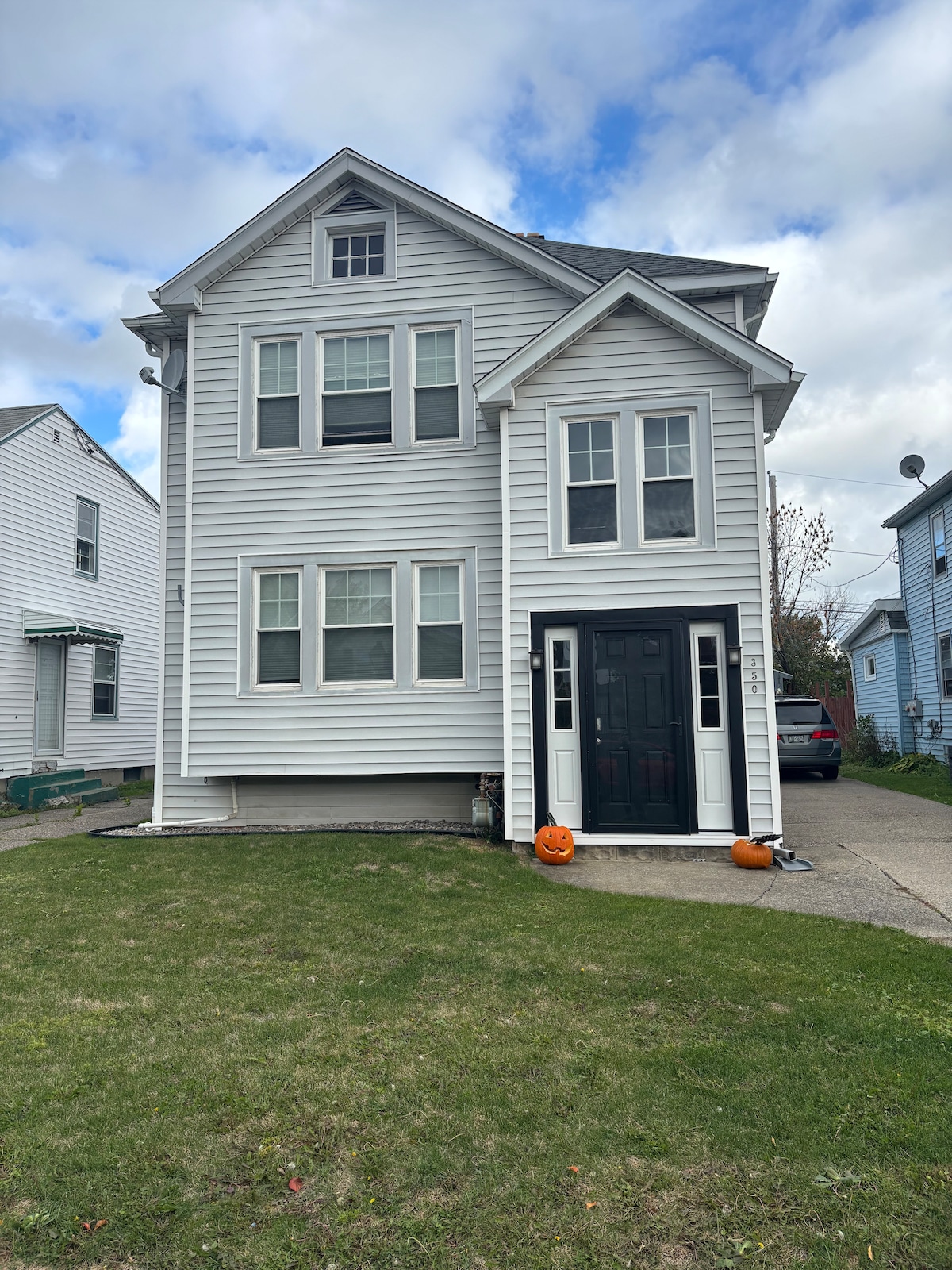The exterior of a three-story house is featured, showcasing a white facade with multiple windows. A black front door creates a contrast at the entrance. Two pumpkins are placed on the ground by the door, and a grassy area surrounds the property.
