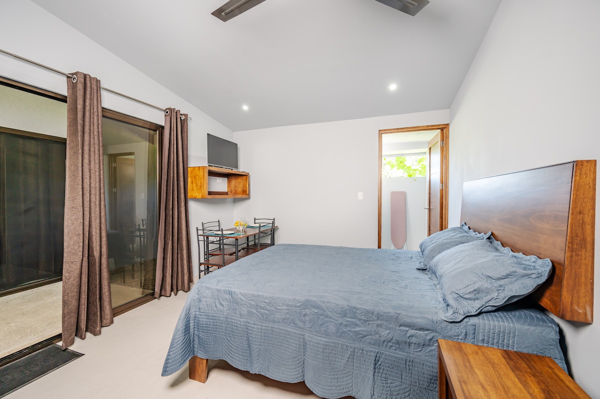 A spacious studio apartment features a comfortable bed with a textured blue coverlet. A small dining area, consisting of a table and chairs, is situated beside sliding glass doors that lead to an outdoor space. Additional natural light is provided through a nearby window.