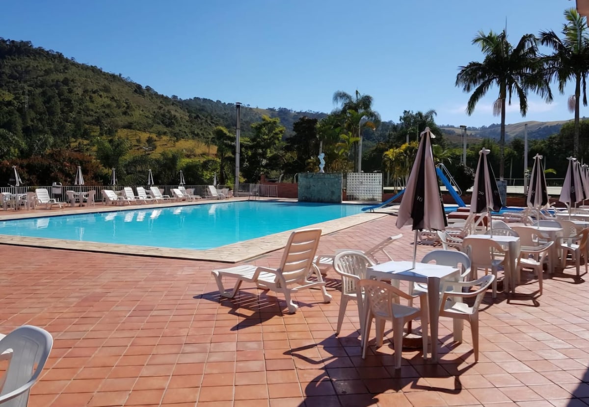 An outdoor pool area is depicted, featuring a large swimming pool surrounded by sun loungers and tables. Umbrellas provide shade, and palm trees line the perimeter. Scenic hills are visible in the background under a clear blue sky.