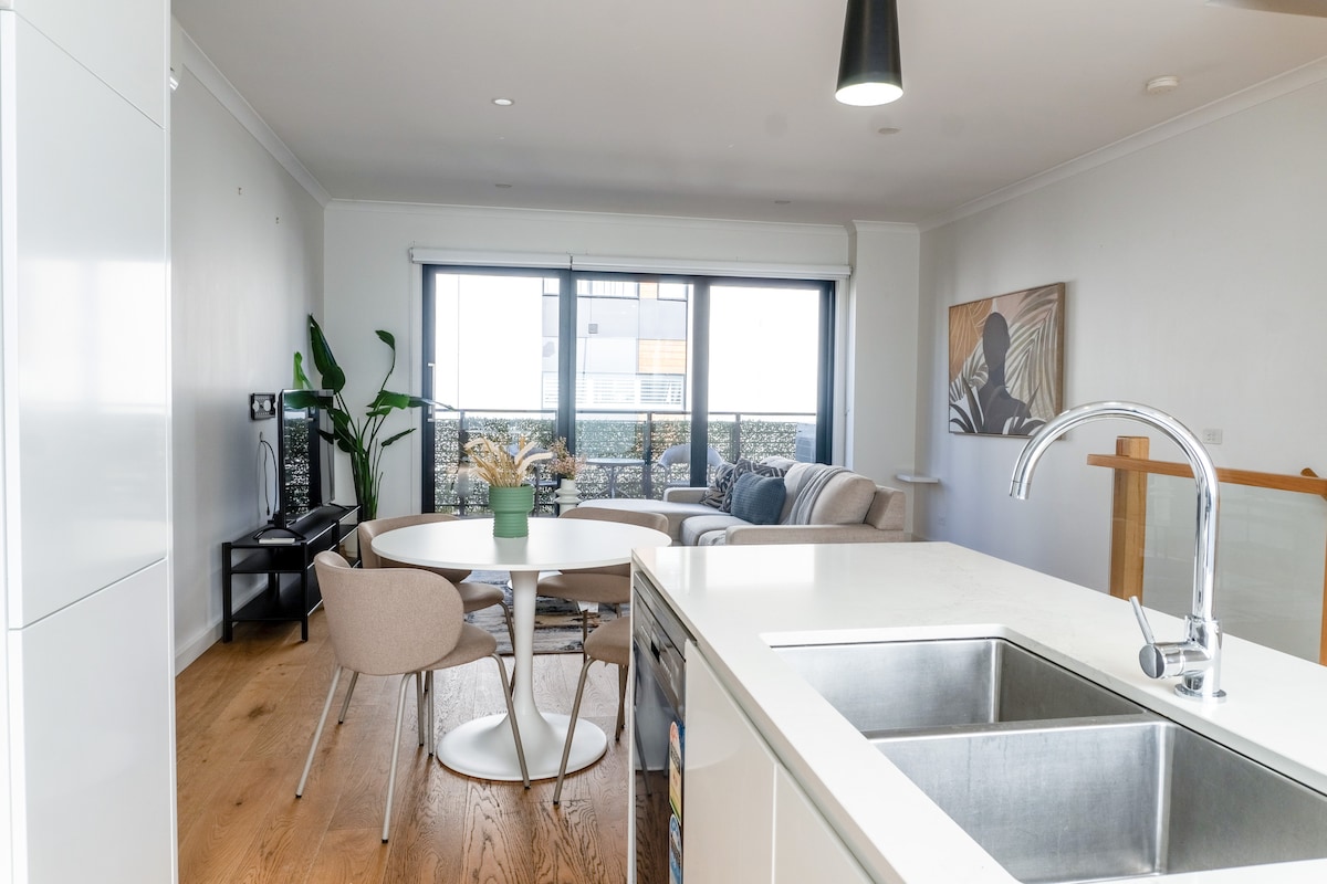 A bright and airy living space is showcased, featuring a dining table with four chairs and a comfortable sofa. Large windows allow natural light to fill the room, while a potted plant adds a touch of greenery. An open kitchen area is visible in the foreground.