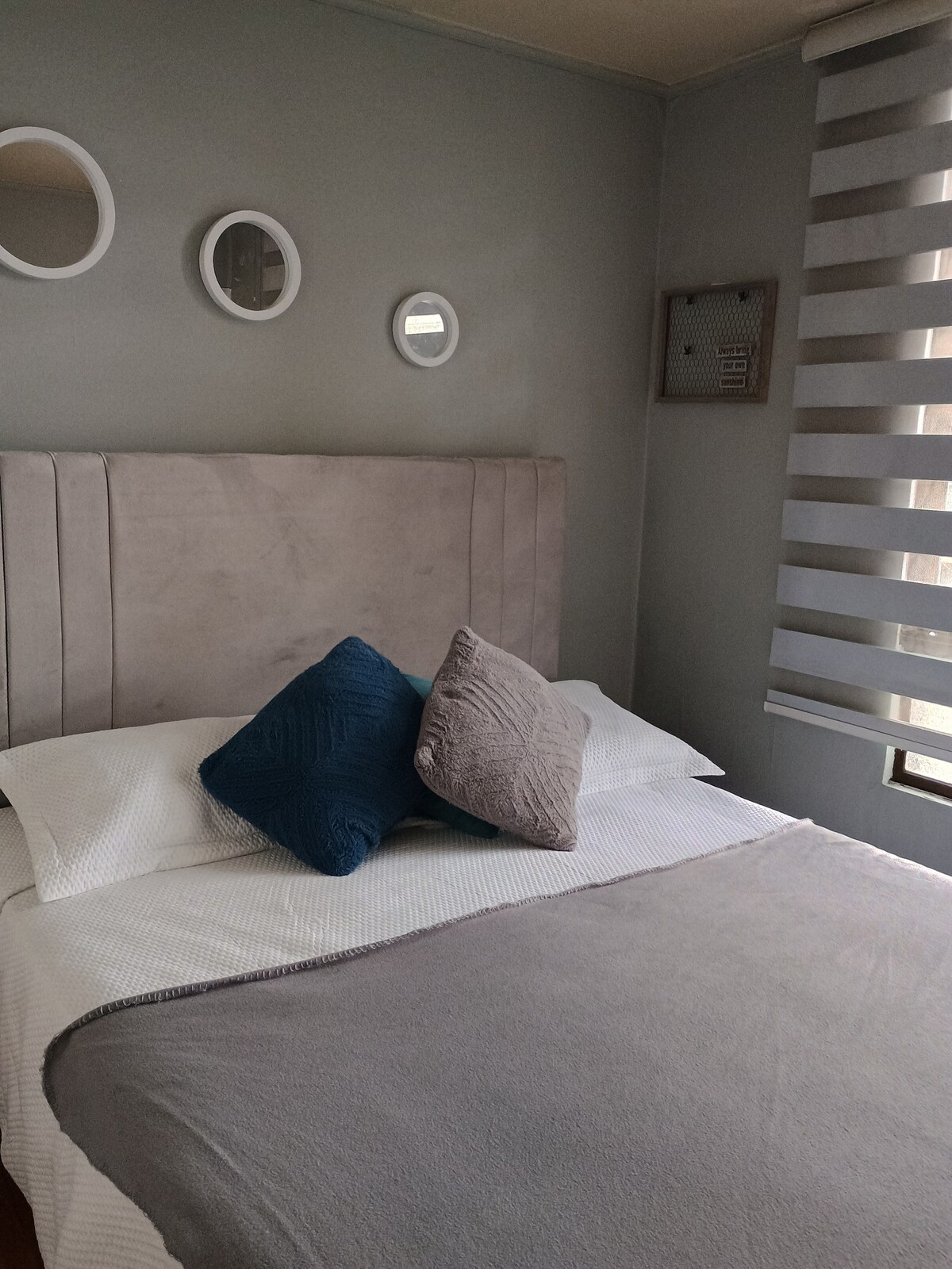 A softly colored bedroom features a spacious bed with a light-colored duvet. Decorative pillows in blue and grey are neatly arranged at the head of the bed. Circular mirrors are mounted on the wall above, while light-filtering blinds provide a gentle illumination.