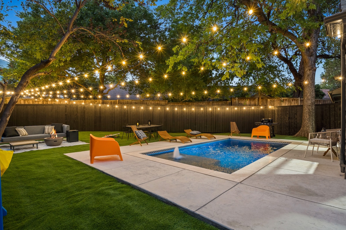 An outdoor space features a private heated pool surrounded by a paved patio, accentuated by lounge chairs and a cozy seating area. Soft glowing lights are strung overhead, enhancing the inviting atmosphere of the spacious yard adorned with greenery.