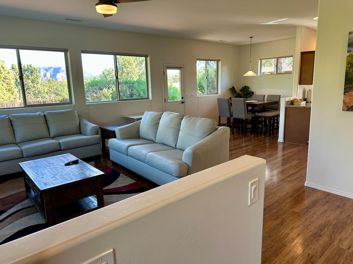 An open-plan living area is highlighted by a light-colored sectional sofa facing a wooden coffee table. Natural light fills the space through multiple windows, offering views of green trees and distant red rock formations. A dining area with a table and chairs is also visible.