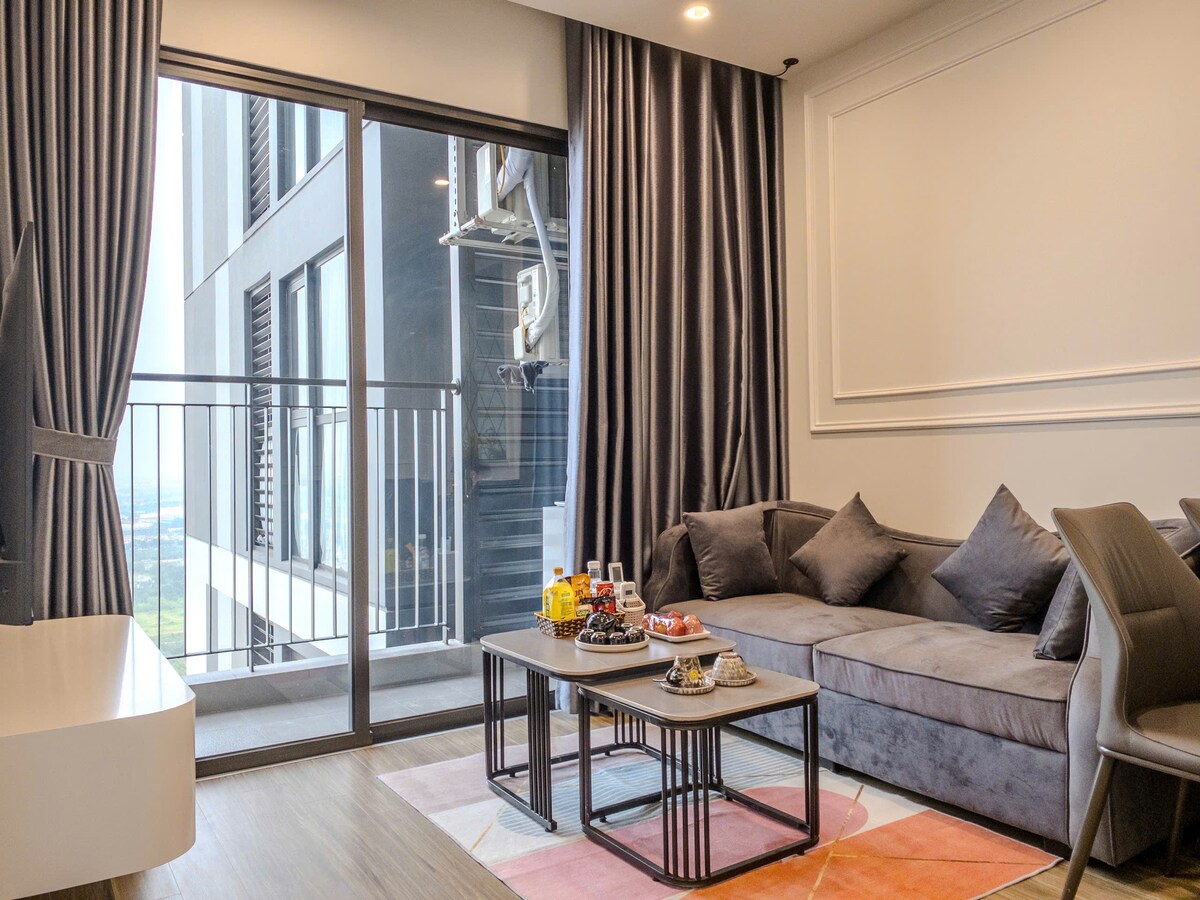 A modern living room features a comfortable grey sofa paired with dark accents. A low coffee table holds various refreshments. Large glass doors open to a balcony, allowing natural light to brighten the space. Plush curtains frame the windows, enhancing the room's contemporary aesthetic.