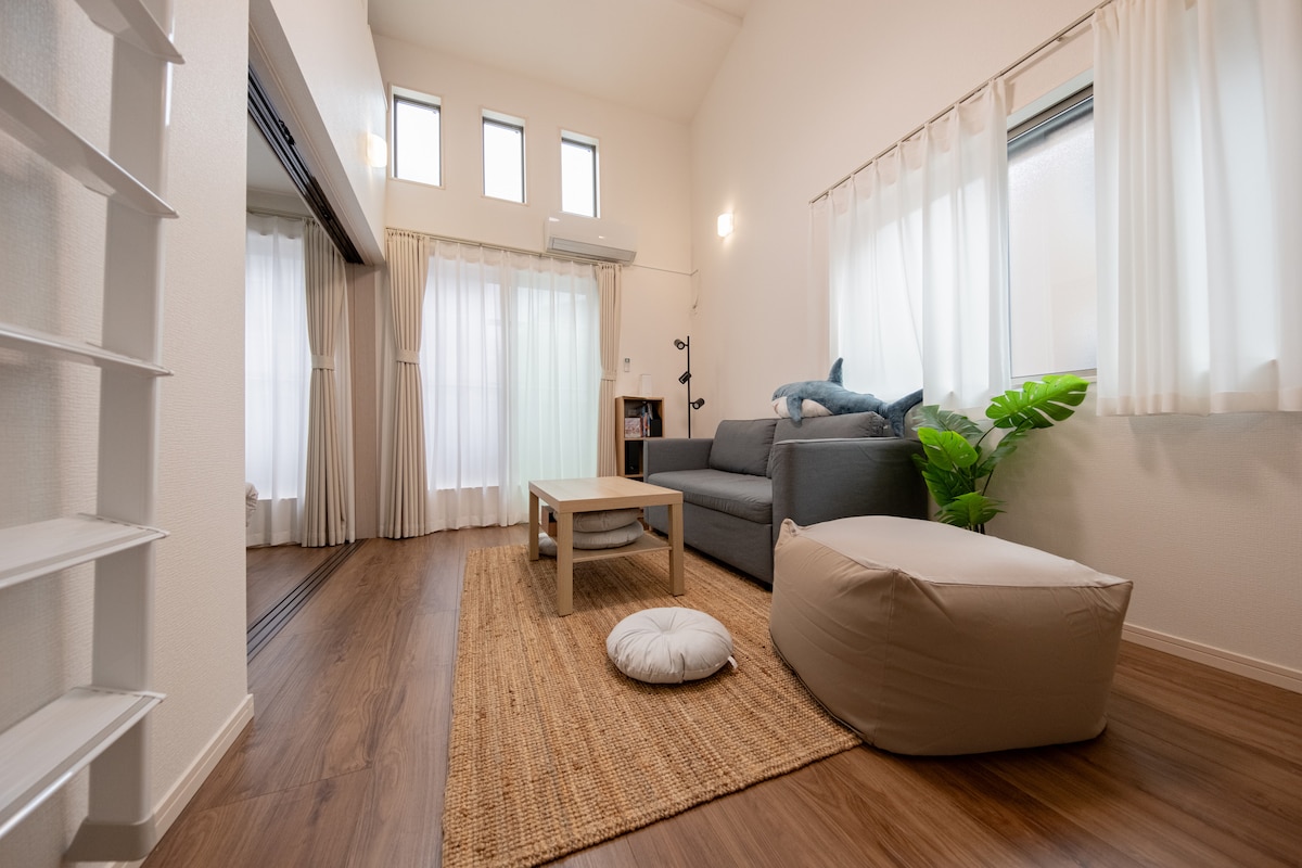 A bright living area features large windows allowing natural light to fill the space. A comfortable gray sofa is arranged with a small coffee table, complemented by a soft floor cushion and a decorative green plant. Natural wood flooring adds warmth to the ambiance.