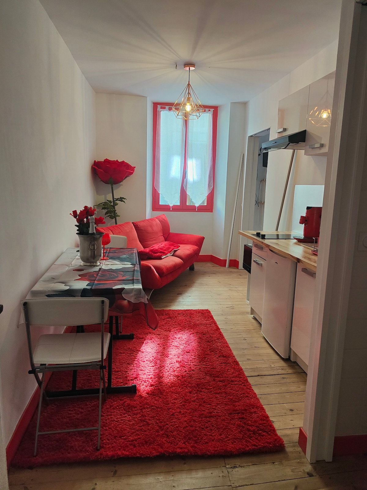 A bright living area is shown, featuring a red sofa and a glass dining table set for two. A vibrant area rug covers the wooden floor, while red accents adorn the walls and windows. Natural light filters through sheer curtains, enhancing the cozy ambiance.