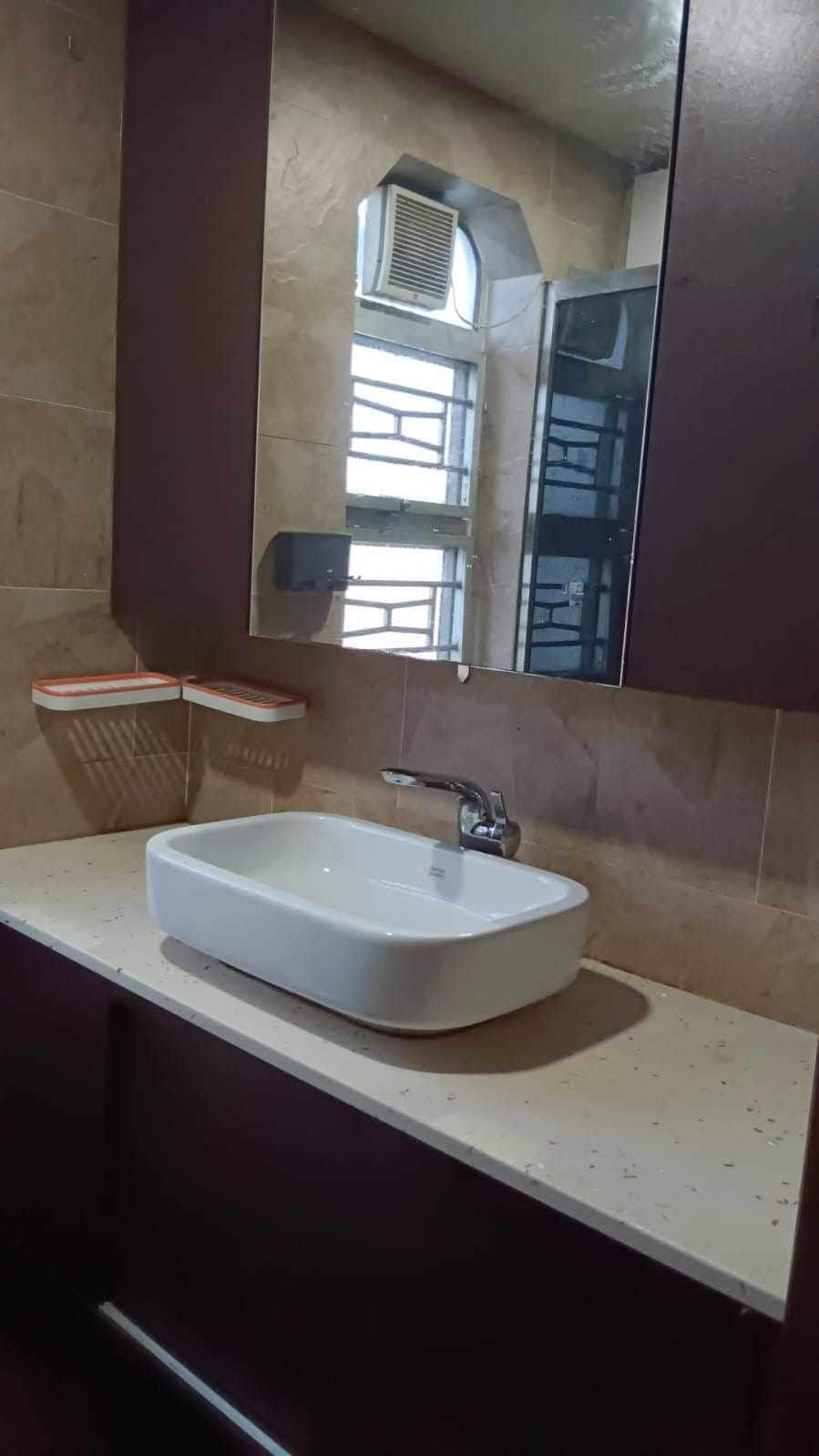 A modern bathroom features a sleek, rectangular basin positioned on a light-colored countertop. The wall-mounted mirror reflects the space, and two small shelves provide storage. Natural light enters through a window, enhancing the neutral tones of the tiled walls.