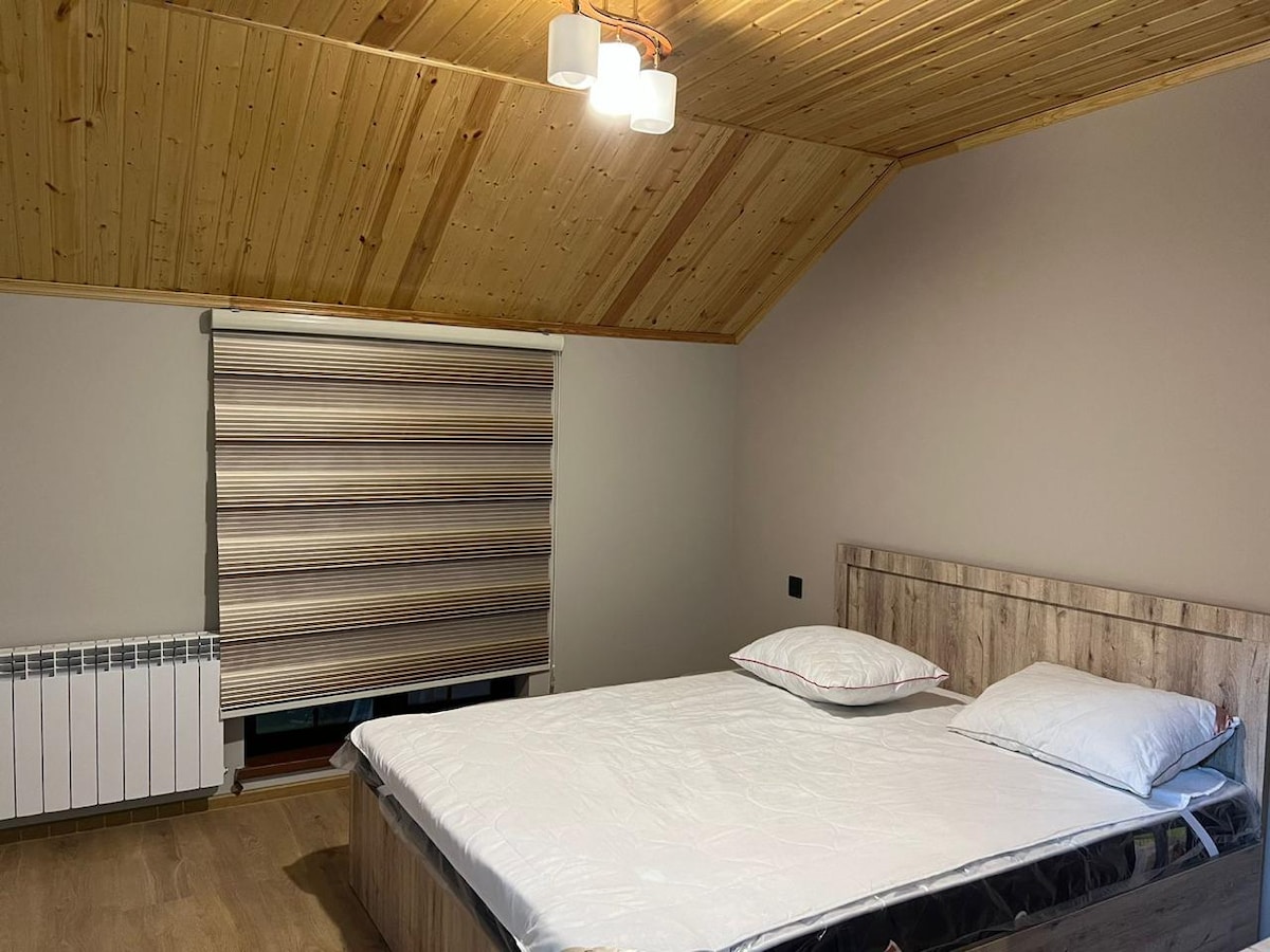 A spacious sleeping area is highlighted by a wooden ceiling and minimalistic decor. A large bed with a simple headboard occupies the center, accompanied by two neatly arranged pillows. A window with rolled shades provides natural light while maintaining privacy.