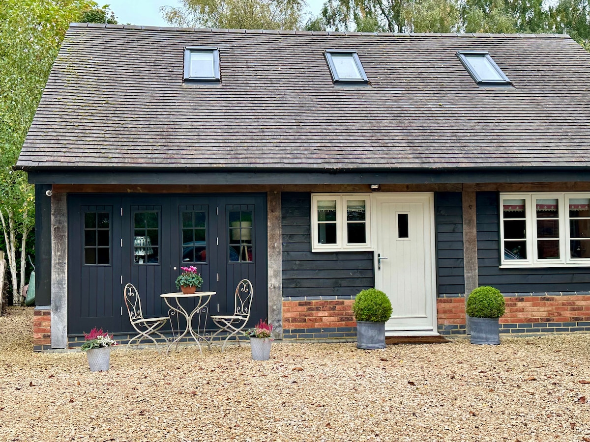 A charming exterior view of The Gallery features a wooden facade with black siding and a welcoming cream door. Two windows and a well-kept gravel area are visible. A small outdoor dining set sits invitingly in front, surrounded by neatly arranged potted plants.