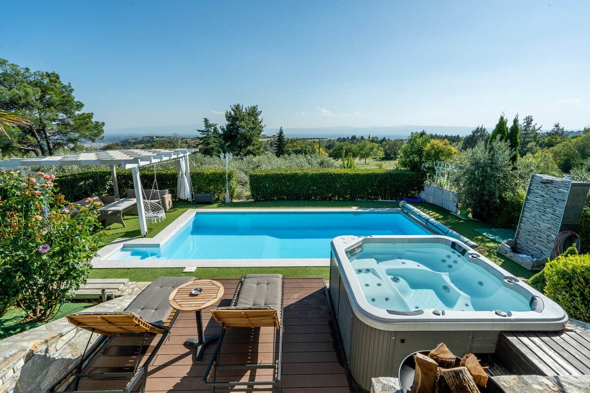 An outdoor area features a private swimming pool and a jacuzzi, surrounded by greenery. Sun loungers are placed nearby with a small table. A pergola offers shade, enhancing the serene view of the landscape and distant hills.