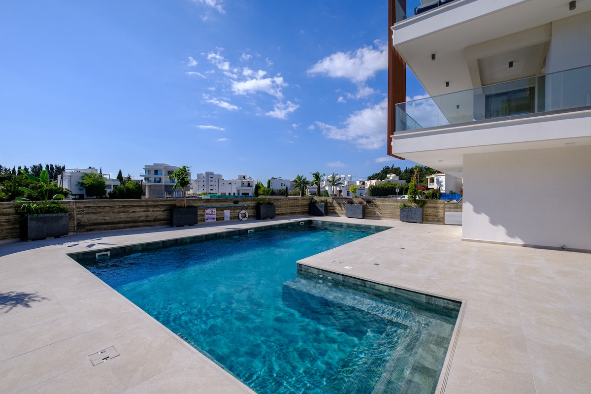 A spacious outdoor pool is surrounded by a modern tiled deck, featuring lounge areas and landscaped greenery. Clear blue water reflects the sky, creating a serene atmosphere. The pool is bordered by a contemporary building with large glass balconies, adding a touch of elegance to the setting.