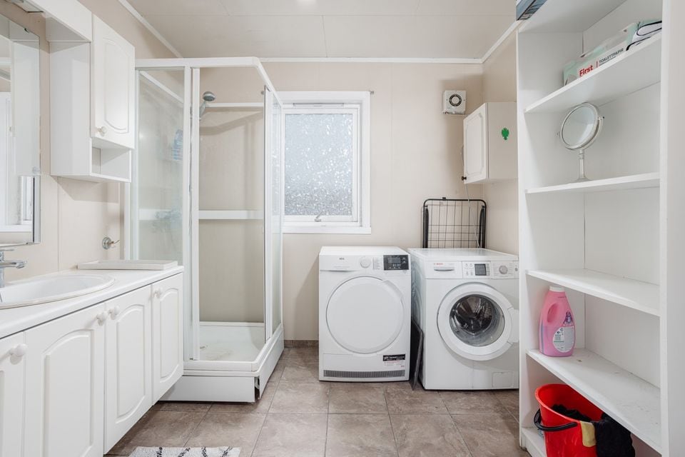 A functional laundry area is equipped with a washing machine and dryer. A glass shower enclosure occupies one corner, while a window allows natural light to enter. Light-colored cabinetry and shelving provide storage options, and the tiled floor is practical for the space.