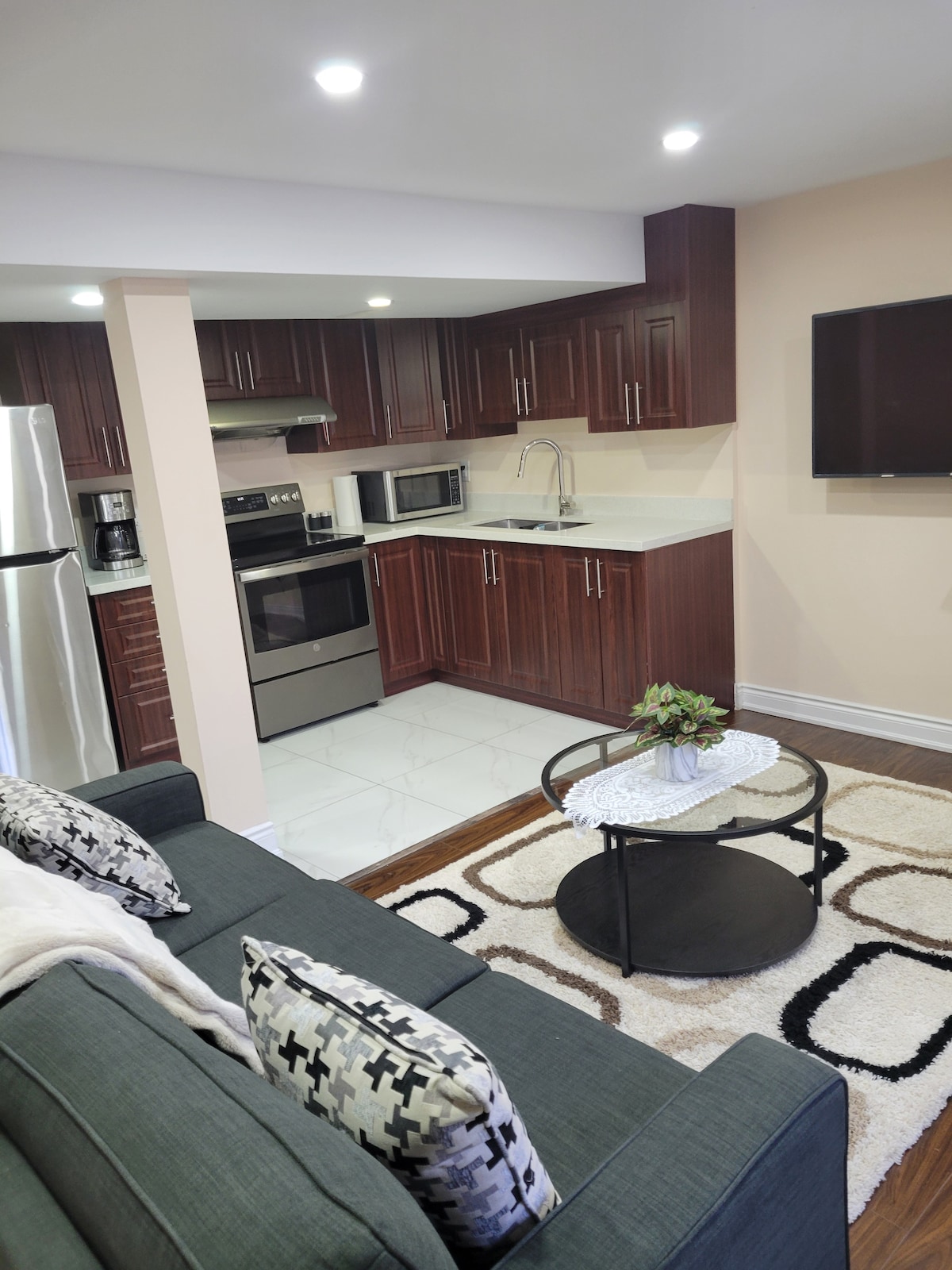 A modern kitchen features dark wood cabinetry with stainless steel appliances, including a refrigerator and stove. A cozy living area is visible, furnished with a gray sofa and a round coffee table on a patterned rug. Kitchen essentials are present, contributing to a functional space.