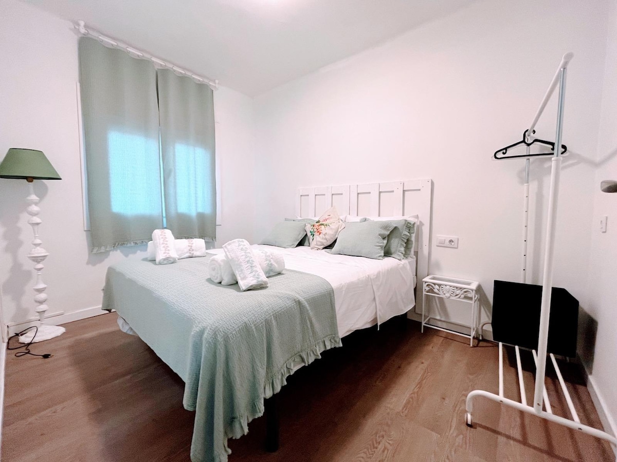 A light and airy bedroom is furnished with a spacious bed dressed in soft green linens. Natural light enters through sheer curtains, illuminating the space. A simple floor lamp, a clothing rack, and a small nightstand are positioned alongside the bed.